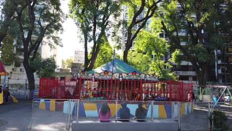 Merry-go-round-kids-playground-at-public-city-park,-greenery-trees-and-kids-at-buenos-aires-argentina,-plaza-flores