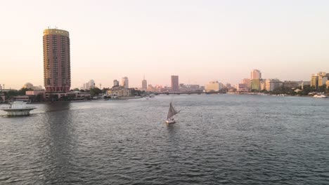 Antena-Sobre-El-Río-Nilo,-Un-Velero-En-El-Agua-Con-La-Torre-De-El-Cairo-Y-El-Hotel-Sofitel-Cairo-Nile-El-Gezirah-Al-Fondo,-En-El-Cairo,-Egipto