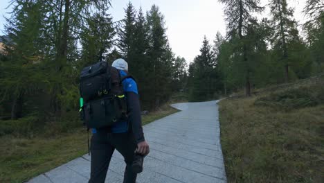 Male-Hiking-Up-Path-In-Forest-With-Backpack-Holding-Camera-In-Valmalenco-Region,-Italy