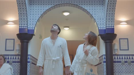 Una-Pareja-Joven-Con-Albornoces-Blancos-En-Los-Pasillos-De-Un-Spa-De-Balneoterapia,-Mirando-Al-Techo-Y-Al-Pozo-De-Luz,-Decorado-En-Blanco-Y-Azul-Con-Mosaicos.