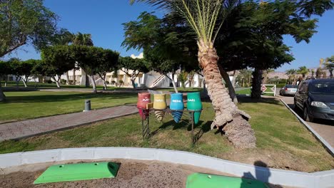 Waste-sorting-in-dahab,-egypt-resort-scene-unfolds-under-a-clear-blue-sky,-featuring-a-palm-tree,-manicured-lawns,-a-walkway-lined-with-trimmed-trees,-colorful-recycling-bins,-a-parked-car