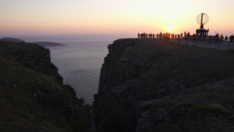 Aerial-View-of-North-Cape-Norway-and-Tourists-Around-Globe-Monument-With-Sunset-Above-Sea,-Drone-Shot