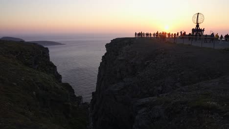 Vista-Aérea-Del-Cabo-Norte-De-Noruega-Y-Turistas-Alrededor-Del-Monumento-Mundial-Con-Puesta-De-Sol-Sobre-El-Mar,-Disparo-De-Drones