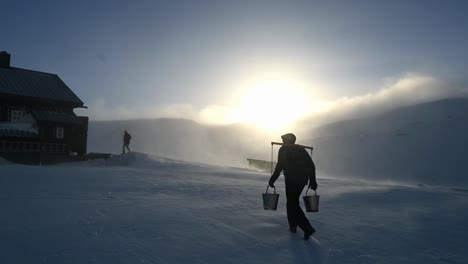 Silhouette-of-man-carrying-two-buckets-of-water-on-shoulder-pole-up-snowy-mountain-towards-cabin