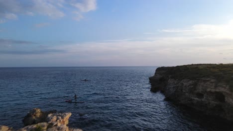 Stand-up-paddle-boarder-going-around-rocks-at-Binigaus-Beach-in-Menorca-at-sunset