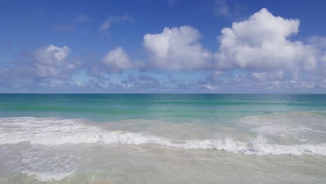 drone-footage-starting-on-a-white-sandy-beach-across-the-turquoise-water-of-Waimanalo-Bay-with-bright-blue-sky-and-puffy-white-clouds