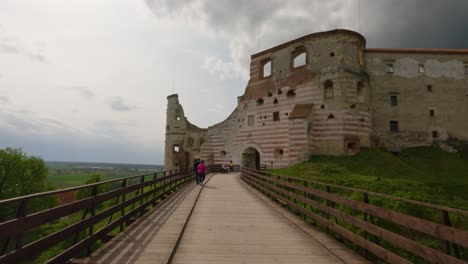 Elder-couple-walk-to-Bolczów-Janowiec-Old-historic-Castle-in-Poland-mountain-side-medieval-architecture-with-cloudy-skyline-and-river-background