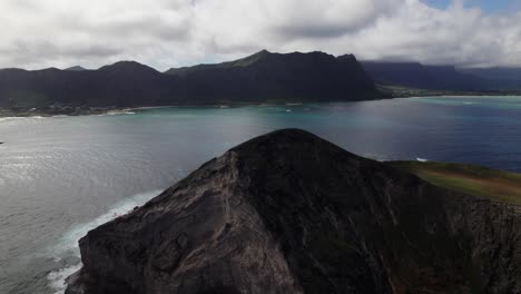 Vista-Aérea-Que-Gira-Alrededor-De-Una-Pequeña-Isla-Montañosa-Frente-A-La-Costa-De-Oahu,-Hawaii,-Comenzando-En-El-Lado-Sombreado-Y-Girando-Para-Mostrar-El-Lado-Iluminado-Por-El-Sol-De-La-Formación-Volcánica.