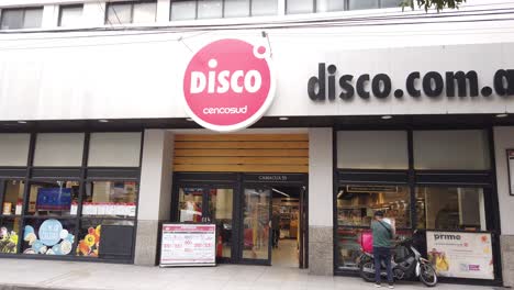 Establishing-shot-at-Disco-supermarket-chain-store-retail-in-Argentina-groceries-shop-people-enter-entrance-door,-cars-traffic-drive-by-Capital-city-of-Buenos-aires