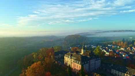 Aerial-View-of-Castle-and-Village-in-Scenic-Landscape-of-Germany-on-Sunny-Misty-Autumn-Morning,-Drone-Shot