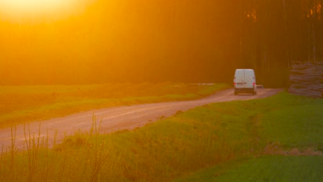 Cars-drive-on-dusty-countryside-gravel-road-during-bright-sunset,-Latvia