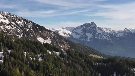 Stunning-drone-shot-of-Amden-Arvenbuel,-Switzerland,-showing-majestic-snow-capped-mountain-ranges-and-picturesque-canopies-of-alpine-trees-with-blue-skies-and-white-clouds-in-a-winter-day