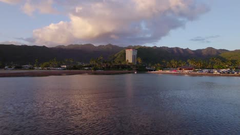 drone-footage-coming-across-the-ocean-at-East-Honolulu-showing-one-tall-building-on-the-edge-of-the-mountain-ridge-in-contrast-to-the-single-story-infrastructure-surrounding-it-pink-alpenglow