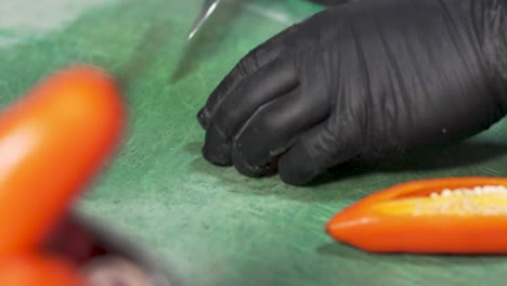 Koch,-Trägt-Eine-Blaue-Jeansschürze-Und-Schwarze-Sterile-Handschuhe,-Schneidet-Und-Schneidet-Gekonnt-Frische-Peruanische-Orange-Chilischoten-In-Streifen-Auf-Einem-Grünen-Kunststoff-Schneidebrett-Mit-Einem-Scharfen-Kochmesser