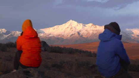 Two-people-in-orange-and-blue-jackets-sit-facing-away-from-each-other-as-alpen-glow-sets-on-Sichuan-Yala-snowy-mountain