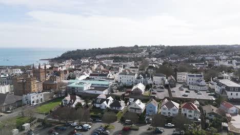 Flight-over-roof-tops-of-St-Peter-Port-Guernsey-towards-Elizabeth-College-with-sea-views-and-wooded-headland-beyond-on-bright-day
