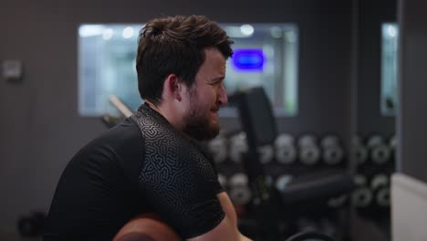 Un-Hombre-Barbudo-Y-Determinado-Realiza-Curl-De-Bíceps-Con-Barra-Sentado-En-El-Gimnasio-Interior