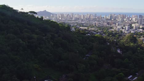 Toma-Panorámica-Hacia-La-Derecha-De-La-Ciudad-De-Honolulu,-Hawaii,-En-La-Isla-De-Oahu,-Con-Una-Exuberante-Ladera-Sombreada-De-Verde-En-Primer-Plano-Y-Los-Altos-Edificios-De-La-Metrópoli-Contra-El-Océano-Al-Final-De-La-Tarde.