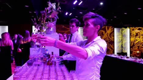 bar-service-during-a-student-gala-evening-with-the-barmaid-serving-two-glasses-of-champagne-to-a-couple-of-young-students,-dark-and-pink-subdued-light,-many-filled-glasses-in-the-foreground