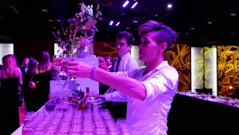 Servicio-De-Bar-Durante-Una-Velada-De-Gala-Estudiantil-Con-La-Camarera-Sirviendo-Dos-Copas-De-Champán-A-Un-Par-De-Jóvenes-Estudiantes,-Luz-Tenue-Oscura-Y-Rosada,-Muchas-Copas-Llenas-En-Primer-Plano