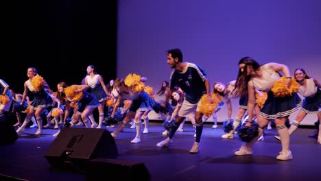 cheerleading-group-with-two-boys-doing-a-choreography-on-a-stage-during-a-gala-evening,-dressed-in-blue-and-white-with-yellow-and-blue-pom-poms