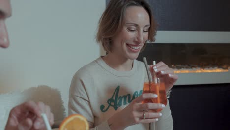 Una-Hermosa-Joven-Caucásica-Sonriente-Alcanzando-Su-Vaso-De-Cóctel-De-Naranja-Para-Brindar-Por-Un-Joven-A-Su-Lado,-Bebiendo-Con-Una-Pajita