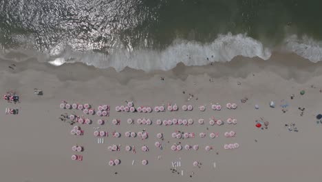 Top-down-shot-of-a-perfect-beach-day-with-uniform-pink-umbrellas-with-the-waves-crashing-on-the-beach