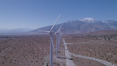 La-Lenta-Panorámica-Del-Dron-Revela-Una-Línea-De-Molinos-De-Viento-En-Funcionamiento-En-El-Desierto,-Con-Montañas-Cubiertas-De-Nieve-Al-Fondo.