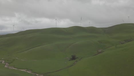 Drone-pulls-out-overlooking-a-large-grassy-hill-with-windmills-on-overcast-day