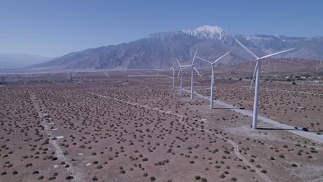 Drone-pulls-back,-revealing-windmills-in-desert-with-snow-capped-mountains-in-background