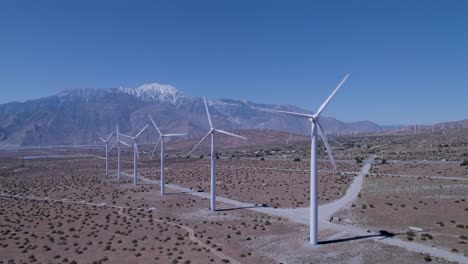 El-Dron-Retrocede,-Revelando-Molinos-De-Viento-En-El-Desierto-Con-Montañas-Cubiertas-De-Nieve-Y-Molinos-De-Viento-Adicionales-En-El-Fondo.