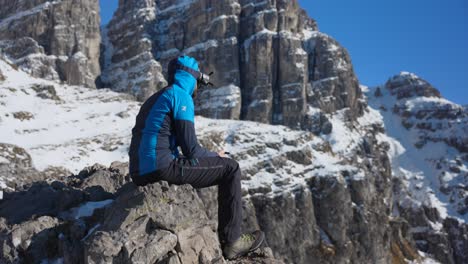 Man-Sitting-On-Mountain-Ledge-Wearing-FPV-Drone-Goggles-Holding-Controller-In-Bergamasque-Prealps