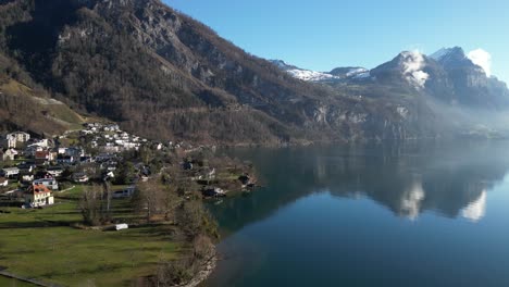 Friedlicher-Drohnenclip,-Der-Einen-Ruhigen-See-Mit-Einem-Dorf-Am-Ufer-Und-Schneebedeckten-Alpenbergen-Im-Hintergrund-An-Einem-Sonnigen-Frühlingstag-Zeigt
