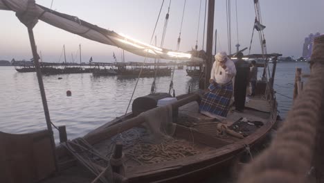 Ein-Emiratischer-Fischer-Sammelt-Muscheln-Und-Perlen-Auf-Einem-Alten-Holzboot-Mit-Der-Flagge-Der-Vereinigten-Arabischen-Emirate-Im-Hintergrund.-Ein-Boot-Liegt-Angedockt-Und-Ein-Fischer-Gewinnt-Perlen-Aus-Muscheln.