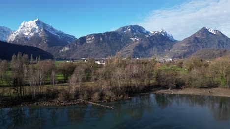 Panning-drone-clip-showing-snowy-mountains-in-Swiss-Alps,-with-grassy-lowlands-and-lake-shore-in-foreground