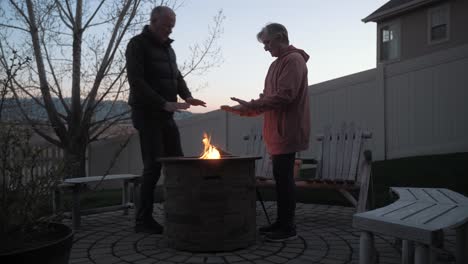 Senior-man-joins-his-wife-as-the-warm-their-hands-at-a-backyard-fire-pit