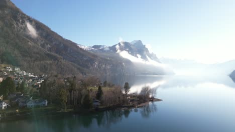 Bright-drone-clip-with-lens-flare-showing-misty-snow-topped-mountains-next-to-calm-lake-in-Switzerland