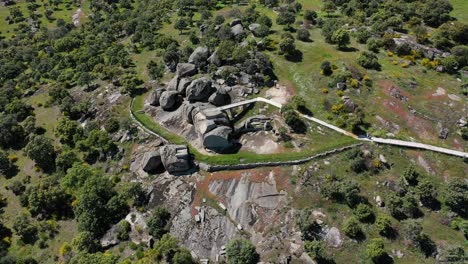 Orbital-flight-with-a-drone-over-a-viewpoint-located-on-a-large-granite-boat-and-large-rocks-around-it,-fenced-in-part-by-a-stone-wall-and-has-a-barbecue-area,-there-are-various-trees-nearby