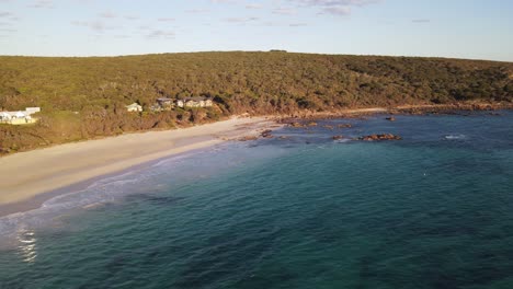 Drone-aerial-over-a-beautiful-blue-beach-next-to-a-national-park-full-of-green-trees