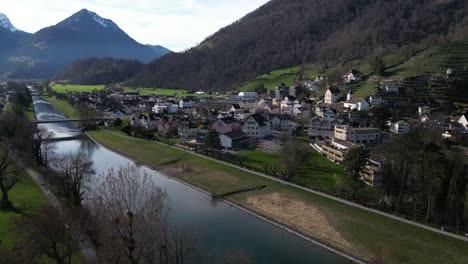 Drohnenclip-Eines-Ruhigen-Flusses-Neben-Einem-Traditionellen-Schweizer-Dorf-In-Den-Alpen