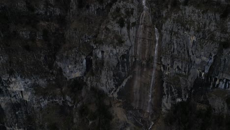 Clip-Ascendente-De-Un-Dron-De-Una-Cascada-De-Alta-Montaña-Que-Muestra-Una-Pared-De-Roca-Empinada-Y-El-Borde-Del-Agua-En-La-Base-Del-Acantilado.