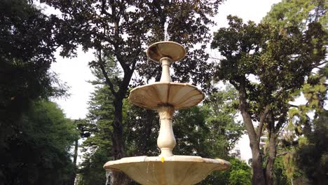 marble-Fountain-water-falling-slow-below-tree-tops-green-dark-skyline-italian-sculpture-panoramic-view-at-natural-environment,-drops-flow