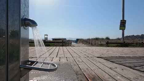 Flowing-drinking-water-from-shiny-stainless-steel-fountain-falling-onto-sea-access-wood-from-village-with-old-wooden-railings,-close-up-blocked-shot,-Pontevedra,-Galicia,-Spain