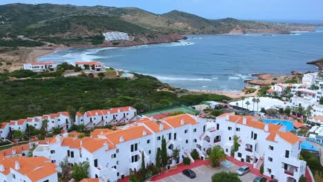 Aerial-views-of-traditional-white-apartments-hotel-with-red-tiled-roofs,-swimming-pool-and-tennis-courts,-with-a-beautiful-sea-cove-in-the-background-in-Minorca,-Spain
