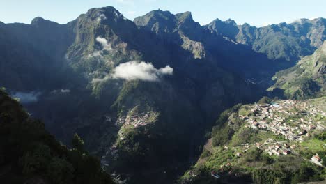 Drone-Shot-of-Small-Village-Located-in-Mountains-Valley,-Covered-by-Shadow-and-White-Clouds