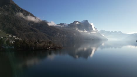 Drone-clip-showing-lake-shoreline-with-misty-and-snowy-mountains-on-bright-sunny-day