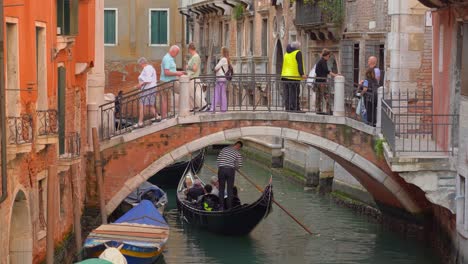 Gondoliers-with-gondolas-in-one-of-many-water-canals-in-Venice-sailing-with-group-of-tourists-beneath-stone-arch-bridge
