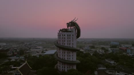 Aerial-closing-up-to-the-captivating-"Dragon-Temple"-Wat-Sam-Phran,-a-remarkable-17-story-cylindrical-pink-temple-adorned-with-a-colossal-green-dragon-encircling-the-structure