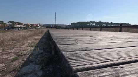 three-people-walk-in-the-distance-on-the-wooden-planks-of-the-overpass-with-old-wooden-railing-on-sandy-soil-in-the-direction-of-the-city,-shot-from-low-turning-right-slow-Pontevedra,-Galicia,-Spain