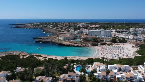 Aerial-shot-of-people-on-vacation-on-beautiful-beach-cove-near-holiday-complex-in-Menorca,-Spain